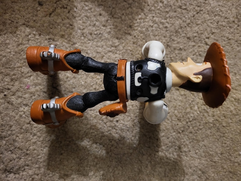 Op de afbeelding: Een plastic action figure van Woody uit de Toy Story-franchise. Woody draagt zijn kenmerkende bruine cowboyhoed, vest en laarzen. Hij ligt op zijn rug met zijn armen gestrekt.