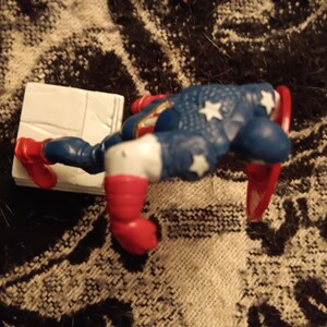 May include: A red, white, and blue action figure of Captain America with a red, white, and blue shield. The figure is posed in a dynamic action pose, with one leg raised and the other leg bent. The figure is standing on a white base.