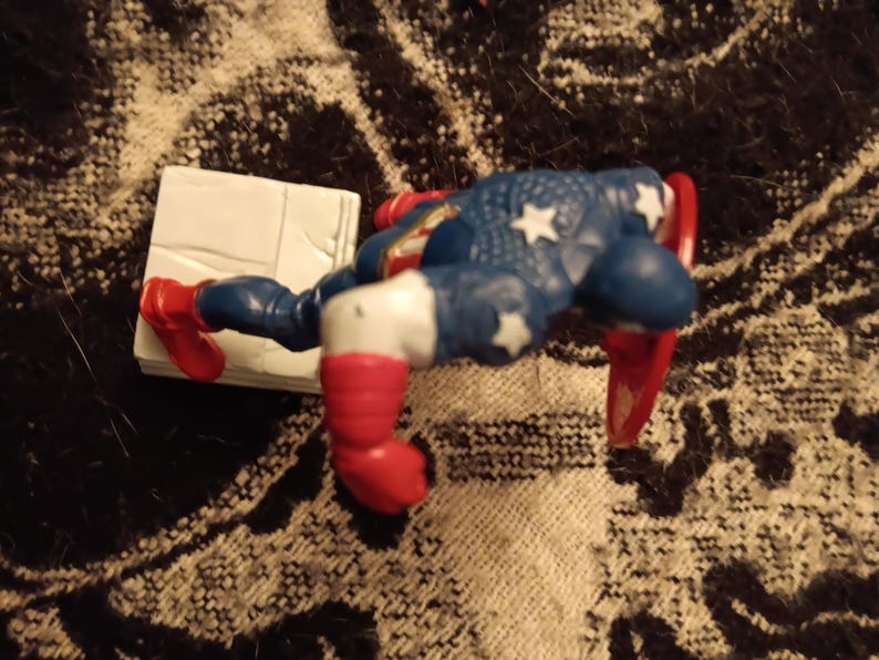 May include: A red, white, and blue action figure of Captain America with a red, white, and blue shield. The figure is posed in a dynamic action pose, with one leg raised and the other leg bent. The figure is standing on a white base.