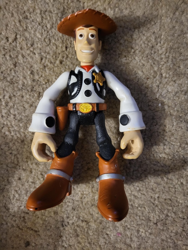 Op de afbeelding: Een plastic speelgoedfiguur van Woody uit de Toy Story-franchise. Woody draagt zijn kenmerkende cowboyhoed, vest en laarzen. Hij heeft een vriendelijke glimlach en staat met zijn armen uitgestrekt.