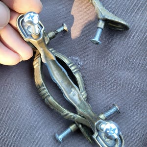 May include: Antique brass drawer pulls with screws. The pulls have an ornate design and are slightly tarnished. The screws are silver. The hardware is likely for a cabinet or dresser. The measurements are in centimetres.