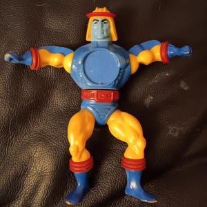 May include: A blue and yellow plastic action figure with a red and yellow helmet. The figure has a blue torso with a circular opening in the center, and yellow legs and arms. The figure is posed with arms outstretched.