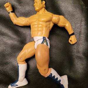以下が含まれることがあります： 白と青のレスリングの衣装を着た黄色のレスリングアクションフィギュア。フィギュアは片手を上げて、もう片方の腕を肘で曲げた状態で走っているポーズをとっている。フィギュアは両方の腕に黒いリストバンドをしている。