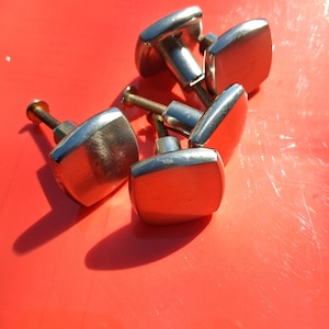 May include: A collection of four square cabinet knobs with a polished silver-tone metallic finish. Each knob features a smooth, slightly curved face and a central threaded metal screw extending from the back for attachment to furniture or doors.