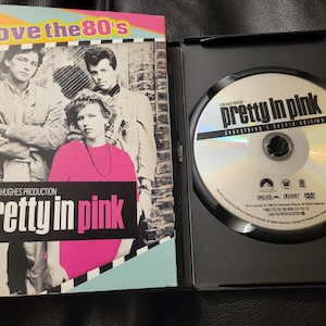 May include: DVD case for the movie "Pretty in Pink." The cover features a photo of the cast with the title in pink and white. The DVD is in an open black case, with the disc visible.