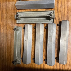 May include: A set of seven silver drawer pulls with black screws. The pulls are rectangular and have a slightly rounded edge. The pulls are arranged in a row on a wooden surface.