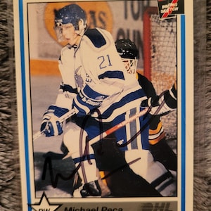 May include: A hockey trading card featuring Michael Peca, a right wing (RW) player for the Erie Otters. The card is from the 1994-95 season of the Ontario Hockey League (OHL). The card is blue and white with a picture of Peca in his hockey uniform. The card has a signature in black ink.