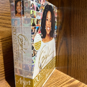 May include: A gold and black DVD box set featuring Oprah Winfrey. The box set is titled "The Oprah Winfrey Show 20th Anniversary Collection" and features a collage of images from the show. The box set is on a wooden surface.
