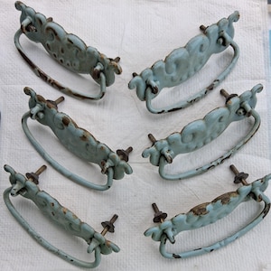 May include: A set of six light blue metal drawer pulls with a distressed finish, featuring ornate scrollwork details and curved drop handles. Each pull includes two threaded metal bolts with nuts for attachment to furniture surfaces.