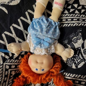 May include: A vintage doll with a plastic head, orange yarn hair, and blue eyes. The doll wears a blue polka-dot dress with a white lace collar and a blue bow. Its legs are covered with beige fabric and white socks with pink bows.