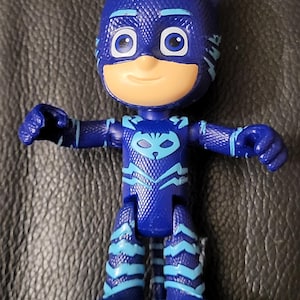 May include: A blue action figure of a character with a cat-like mask and ears. The figure has a light tan face, large blue eyes, and a blue and light blue costume. The figure's arms are outstretched, and it is standing on a dark surface.