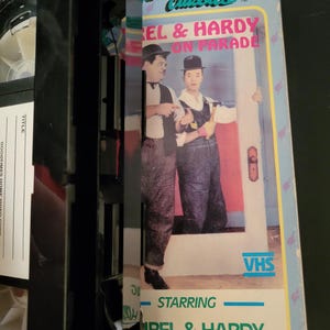 Może przedstawiać: Pudełko kasety VHS z obrazkiem Laurel i Hardy na przodzie. Tytuł kasety to "Laurel & Hardy On Parade" i jest częścią serii "Goodtimes Golden TV Classics".