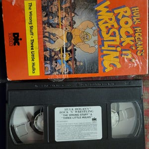 Puede incluir: Una caja de cinta VHS naranja con el título "Hulk Hogan's Rock 'N' Wrestling" e ilustraciones de dibujos animados de luchadores. La cinta VHS negra tiene una etiqueta blanca con los títulos "The Wrong Stuff" y "Three Little Hulks".