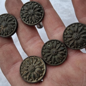 May include: A collection of six antique bronze-colored buttons with a detailed floral design. The buttons are round with a central raised element and radiating petal-like patterns. The buttons are held in a hand, showcasing their aged appearance.