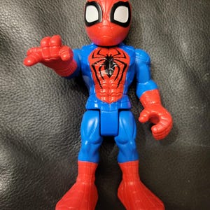 May include: A plastic Spider-Man action figure in a classic pose. The figure features a red mask with white eye lenses, a blue torso and legs, and red hands and feet. A black spider emblem is on the chest.