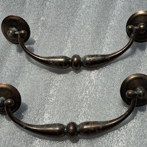 May include: Two matching antique-style brass drawer pulls with a dark, weathered patina. Each handle features a curved, tapered bar with a central decorative knob and two circular mounting plates at the ends, connected by small, articulated joints.