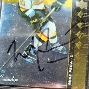 May include: A hockey trading card featuring a player in a yellow, black, and blue uniform, holding a hockey stick. The card has a signature and the text "Canucks" and "MIKE PECA C". The card has a gold border.
