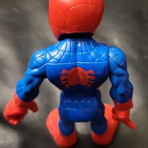 May include: A plastic Spider-Man action figure, viewed from the back. The figure is primarily blue with red accents on the head, hands, and feet. A red spider emblem is on the back. The figure is kneeling.