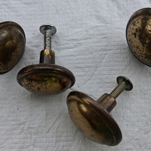 May include: Four round cabinet knobs with a dark, weathered brass finish. Each knob features a smooth, domed top and a threaded metal screw extending from the back for installation. The metal surfaces show signs of patina and uneven dark coloring.