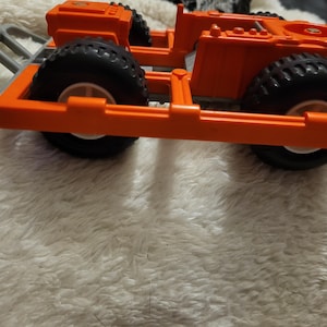 May include: Orange toy truck with black wheels and a gray metal bumper. The truck has a flatbed and is made of plastic.