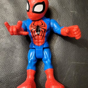 May include: A Spider-Man action figure with a red head and boots, blue torso and arms, and a red chest. The figure has a black spider emblem on the chest and white eye shapes.