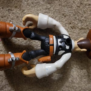 Op de afbeelding: Een plastic speelgoed action figure van een cowboy met een bruine hoed, een zwarte broek, een wit shirt en bruine laarzen. De figuur heeft een zwart-wit vest met een zilveren gesp.
