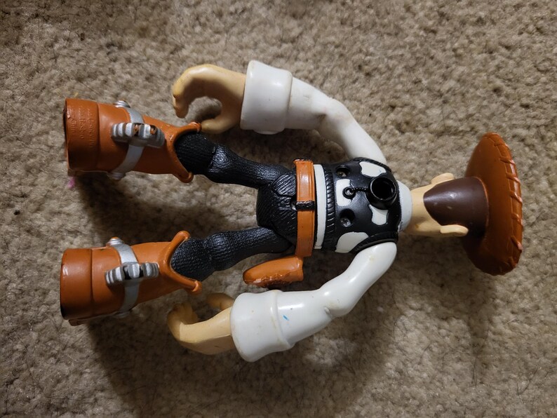 Op de afbeelding: Een plastic speelgoed action figure van een cowboy met een bruine hoed, een zwarte broek, een wit shirt en bruine laarzen. De figuur heeft een zwart-wit vest met een zilveren gesp.