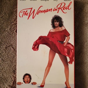 May include: A VHS tape cover for the movie "The Woman in Red" starring Gene Wilder, Charles Grodin, Joseph Bologna, Judith Ivey, and Gilda Radner. The cover features a woman in a red dress with her back to the camera, and a man peeking out from behind a wall.