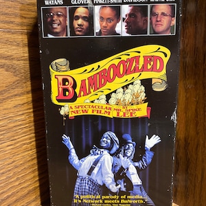 May include: A VHS tape cover for the film "Bamboozled" by Spike Lee. The cover features a yellow and black title card with the movie title in large, white letters. The cover also features a photo of the two main actors, Damon Wayans and Savion Glover, in costume as minstrels. The cover also features a quote from a Time Magazine review of the film.