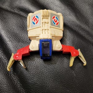May include: A vintage toy robot in off-white, red, blue, and silver. The robot has a rectangular body with two circular designs on the front. It has two red arms with yellow and silver claws. The robot is a collectible toy.