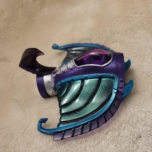 May include: A futuristic, stylized helmet in shades of purple, teal, and silver. The helmet features a curved visor, a rounded top with a purple lens, and intricate detailing. The design suggests a science fiction or fantasy theme.