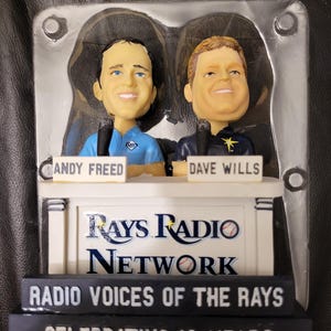 May include: A bobblehead set of two radio announcers, Andy Freed and Dave Wills, in front of a white sign that reads "Rays Radio Network" and "Radio Voices of the Rays Celebrating 10 Years".