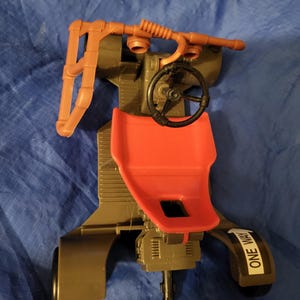 May include: A toy vehicle with a red seat, black steering wheel, and brown piping. The vehicle has a dark green body and the words "ONE WAY" are printed on the side. The toy is made of plastic.