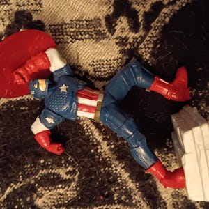 May include: A red, white, and blue action figure of Captain America, wearing his iconic costume and holding his shield. The figure is posed in a dynamic action pose, with one leg raised and the shield held out in front of him. The figure is made of plastic and is approximately 6 inches tall.