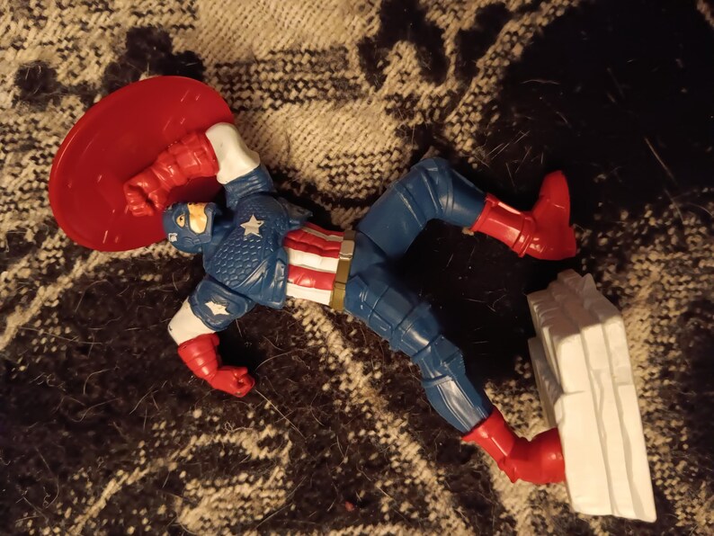 May include: A red, white, and blue action figure of Captain America, wearing his iconic costume and holding his shield. The figure is posed in a dynamic action pose, with one leg raised and the shield held out in front of him. The figure is made of plastic and is approximately 6 inches tall.
