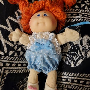 Op de afbeelding: Een vintage pop met oranje garen haar, blauwe ogen en een glimlachende uitdrukking. De pop draagt een blauwe polka-dot outfit met een witte kanten kraag en witte schoenen met roze strikjes. Een klassiek verzamelobject.