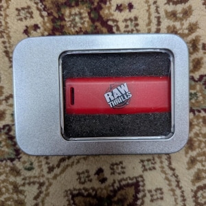 May include: A rectangular silver metal tin with a clear window, containing a red USB flash drive nestled in black foam. The red flash drive features the white text RAW THRILLS printed on its surface, along with a small black rectangular slot on the left.