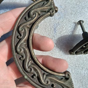 May include: A set of antique-style brass drawer pulls featuring a curved, crescent-shaped design with intricate embossed scrollwork and a dark, weathered patina. The hardware includes a matching smaller knob with a threaded metal mounting screw.
