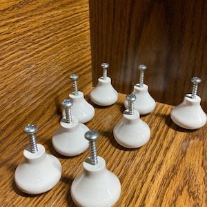 May include: Nine white ceramic cabinet knobs with silver screws. The knobs are shaped like a bell with a rounded top and a slightly flared base.