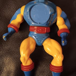 Puede incluir: Una figura de acción de plástico azul y amarillo con un casco rojo y amarillo. La figura tiene una placa de pecho azul con una abertura circular y un cinturón rojo con una hebilla plateada. La figura lleva botas azules con detalles rojos.
