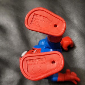 May include: A Spider-Man action figure in a crouching position, viewed from below. The figure is primarily blue and red, with red feet and hands. The soles of the feet are red and bear the Hasbro SA and Marvel & Subs. markings.