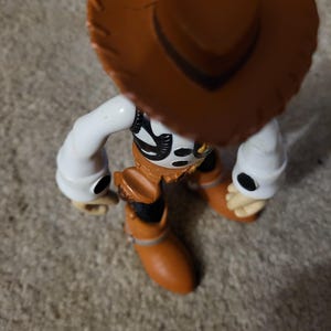 Op de afbeelding: Een bruin en wit plastic speelgoedfiguur van Woody uit Toy Story. Woody draagt een bruine cowboyhoed, een wit shirt, een bruin vest en een bruine broek. Hij houdt een touw vast in zijn rechterhand.