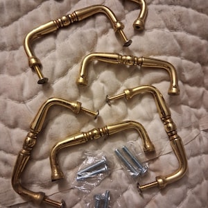 May include: Set of six gold-toned cabinet handles with decorative detailing. Each handle has a curved design with screw attachments. Includes a small package of screws for installation. The handles are arranged on a textured surface.