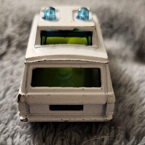 May include: White toy van with blue flashing lights on the roof. The van has a green tinted rear window.