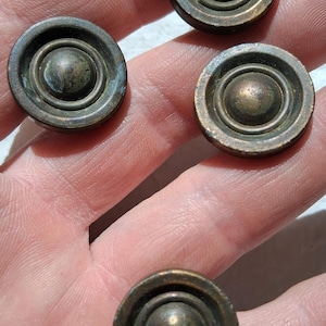 Puede incluir: Cuatro botones metálicos circulares con un acabado de bronce oscuro y desgastado. Cada botón presenta una cúpula central lisa y elevada, rodeada por un anillo circular hundido y un borde exterior plano, con signos de oxidación y pátina.