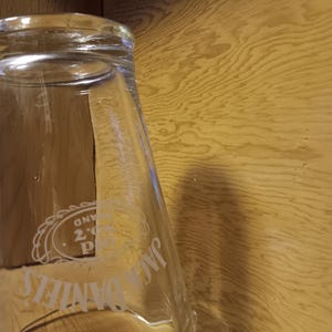 Puede incluir: Un vaso transparente con las palabras "Jack Daniel's" y un logotipo grabado en el lateral. El vaso tiene una boca ancha y un cuerpo cónico. El fondo es una superficie de madera.