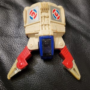 May include: A vintage toy robot with a cream-colored body, red and blue accents, and articulated legs. The robot features circular emblems with red, blue, and white details. The legs are red with yellow claws.