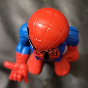 May include: A red and blue Spider-Man action figure. The figure has a red head with a web pattern, blue arms and legs, and red hands and feet. The figure is in a crouching position with one hand extended.