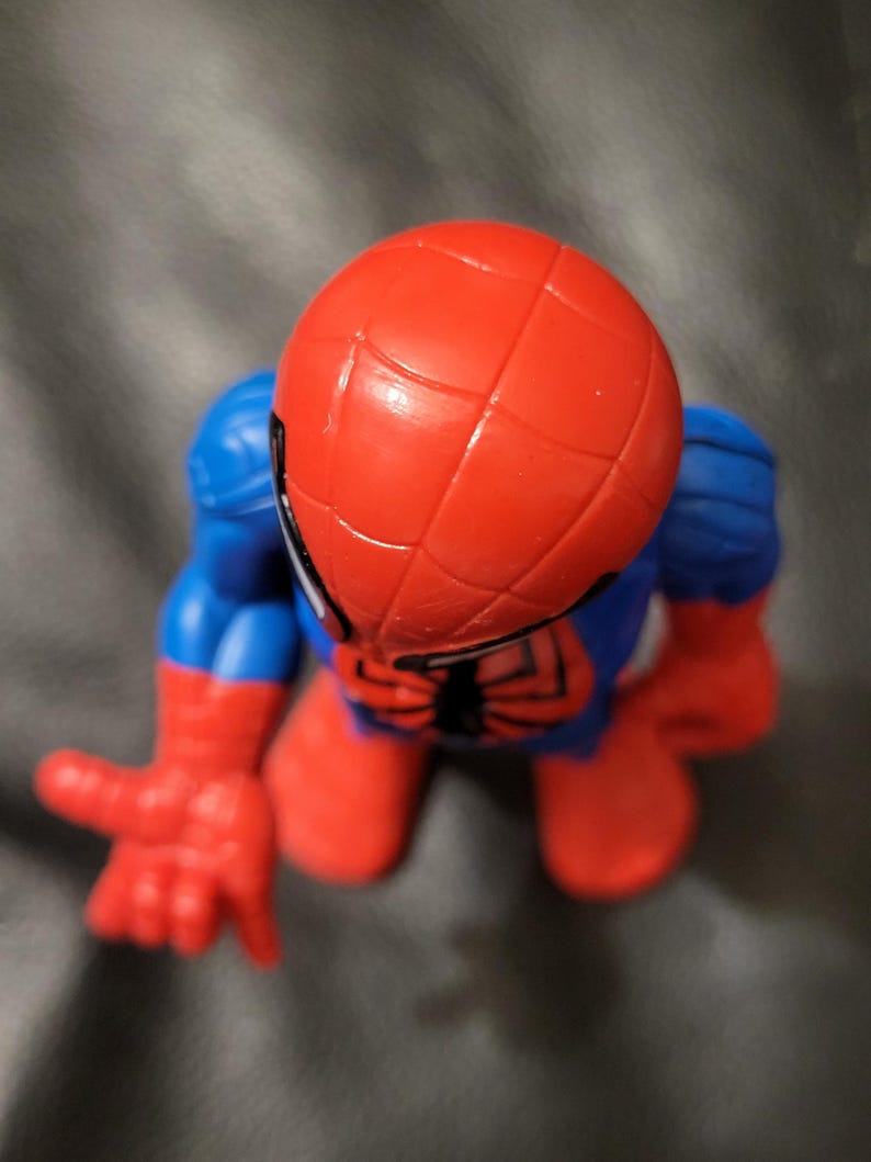 May include: A red and blue Spider-Man action figure. The figure has a red head with a web pattern, blue arms and legs, and red hands and feet. The figure is in a crouching position with one hand extended.
