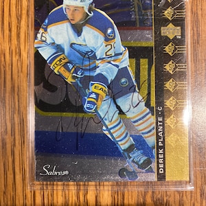 May include: A hockey trading card featuring Derek Plante, a center for the Buffalo Sabres. The card is gold and black with a silver background. The card features a photo of Plante in his hockey uniform, skating with the puck. The card is signed by Plante.